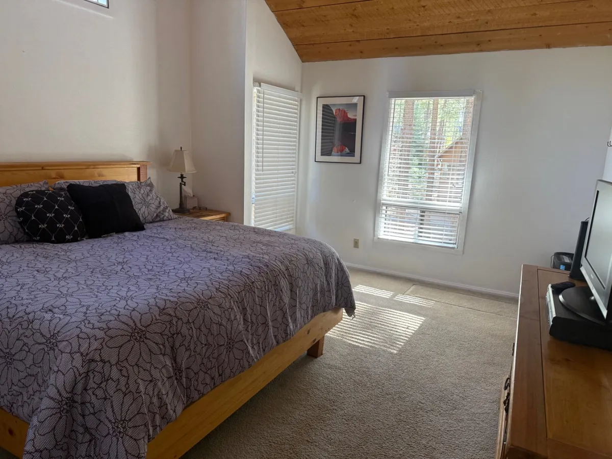A tranquil bedroom featuring a bed adorned with a floral-patterned blanket, warm wooden furniture, and large windows that flood the room with natural light. The space feels peaceful and is ideal for relaxation, reading, or enjoying a morning coffee.