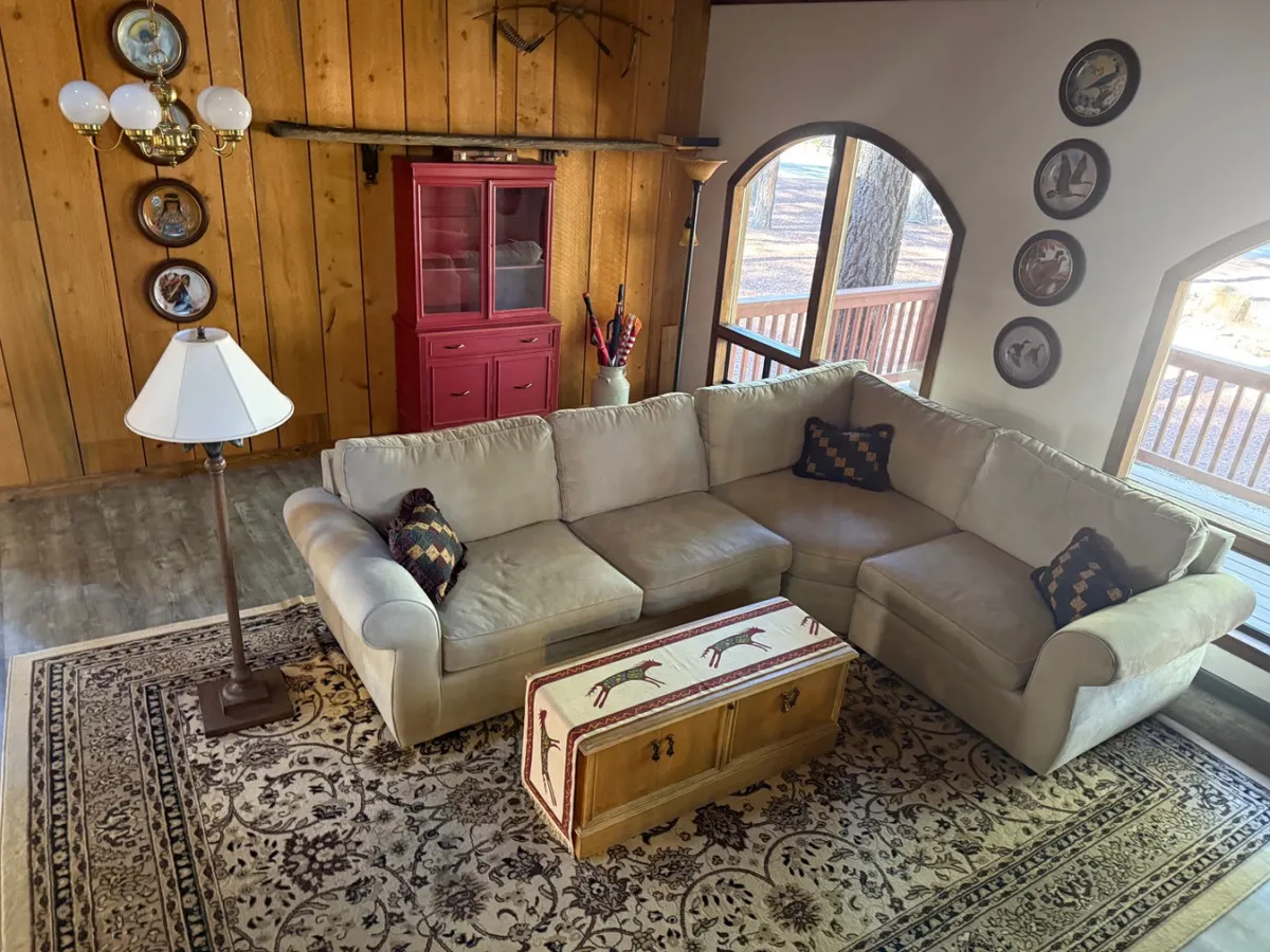 A warm, welcoming living area with a sectional sofa, coffee table, and wall decor, perfect for gathering and conversation. Large windows connect the interior to the outdoors, filling the space with natural light and offering glimpses of the surrounding landscape.