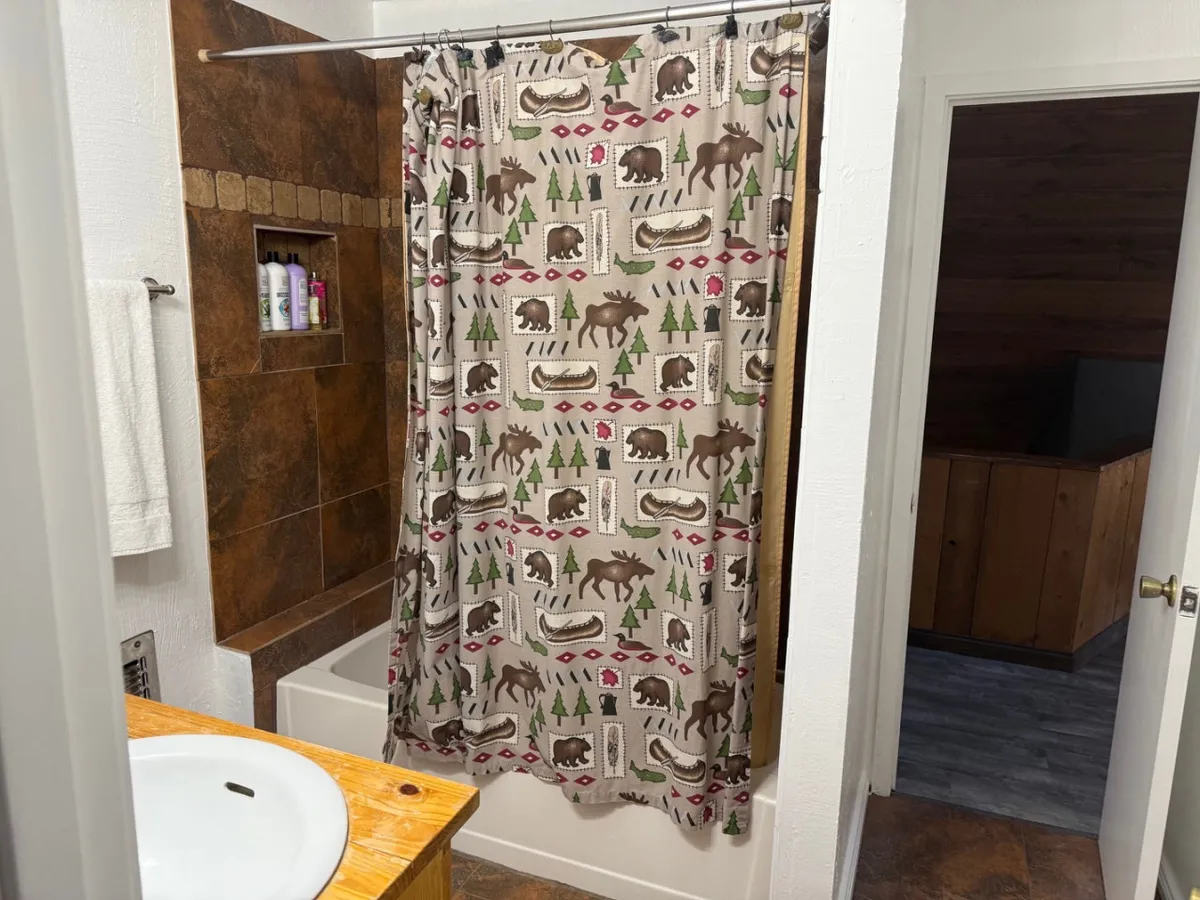 A bathroom with a nature-themed shower curtain, wooden vanity, and tiled wall. The setting is inviting and evokes warmth, offering a sense of relaxation and self-care suited for unwinding after a busy day.