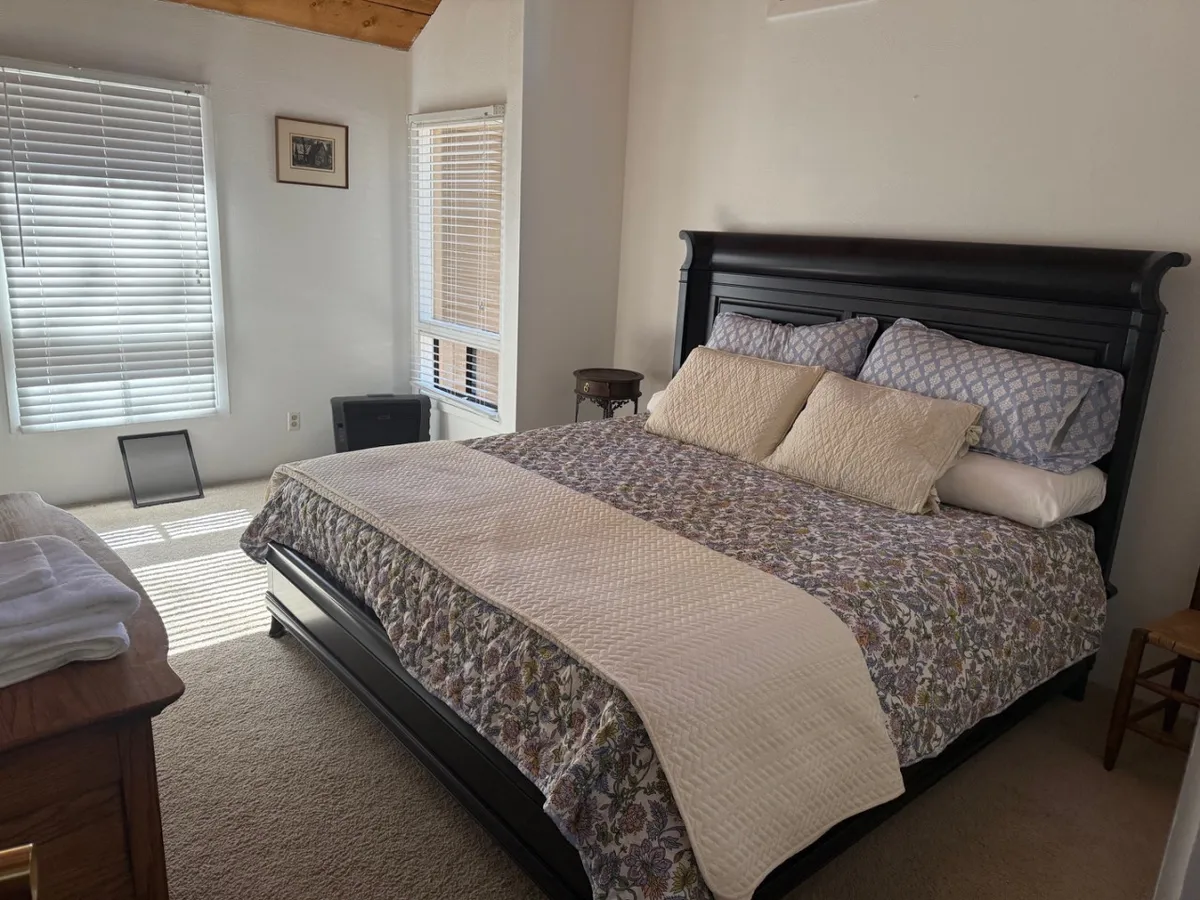 A cozy bedroom scene featuring a neatly made bed positioned beside large windows that let in plenty of natural light, a nightstand with a reading lamp, and a framed picture on the wall. The space feels restful and inviting, offering glimpses of the outdoors and suggesting a relaxing retreat after a day of exploration.