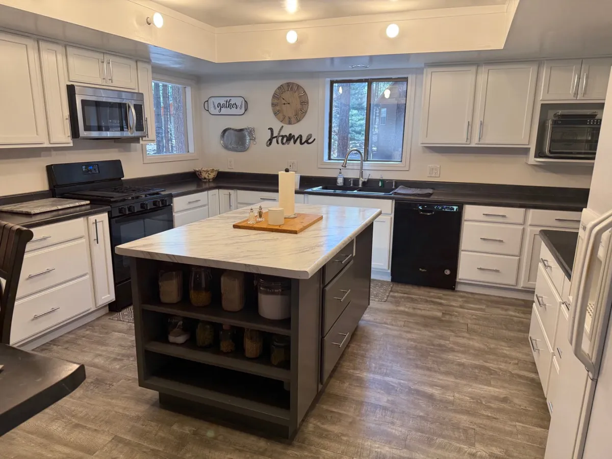 A homey, inviting kitchen scene centered around a spacious island, with a sink and stove visible. Decorative wall signs add character, while large windows let in natural light and offer glimpses of the outdoors. The space is designed for cooking, gathering, and entertaining, embodying the heart of the home.