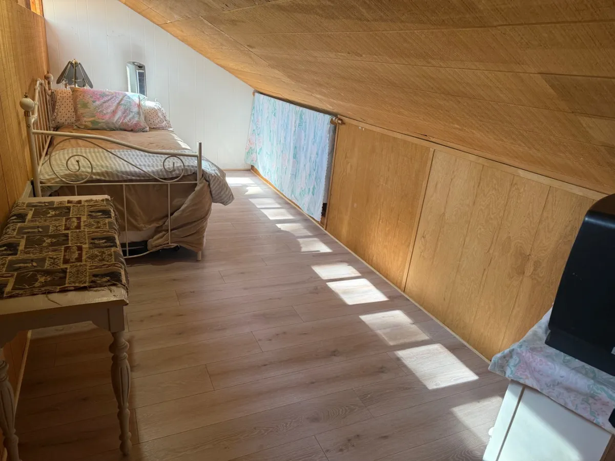 A tranquil corner of a bedroom featuring a bed, a wooden desk positioned near a large window, and warm wood paneling throughout. Sunlight pours into the space, creating an inviting atmosphere perfect for reading, reflecting, or enjoying moments of quiet relaxation.