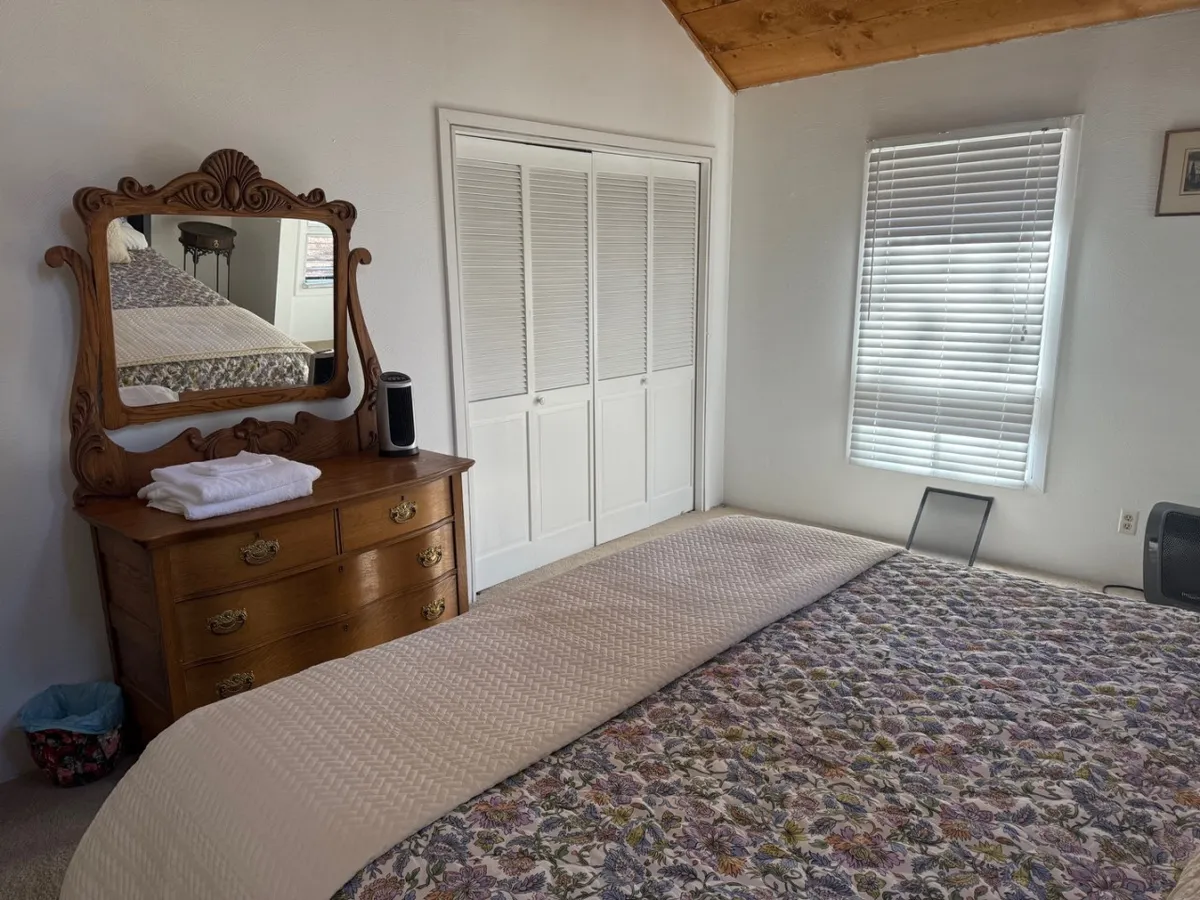 A warmly lit bedroom with floral bedding, an ornate wooden mirror, and a dresser. The scene feels inviting and cozy, ideal for moments of rest or relaxation, with personal touches that evoke comfort and privacy.