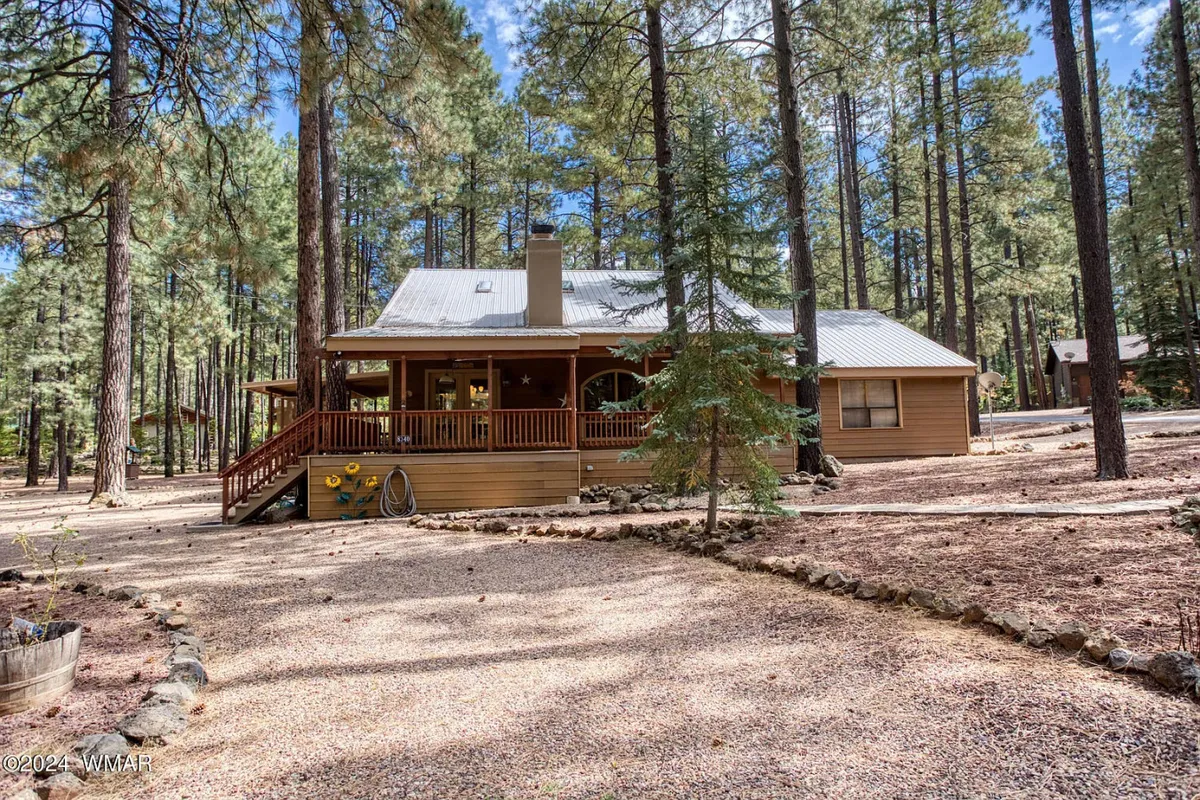 The hero image showcases a charming wooden wraparound porch nestled among towering pine trees, with a gravel driveway leading up to the house. The setting exudes tranquility, inviting guests to relax outdoors, soak in the forest views, and enjoy peaceful moments surrounded by nature.